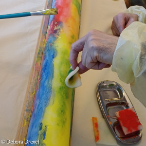 auf einem Tisch liegt eine lange bunte Kerze. rechts hält eine Hand einen Schwamm mit Farbe an die Kerze. Links ist ein Pinsel an der Kerze zu sehen.