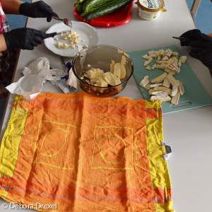Nahaufnahme von einem Tisch auf dem Gemüse und Salat bereitliegt. Ausschnitt von zwei Personen, die gerade Camembert für Obazda schneiden.